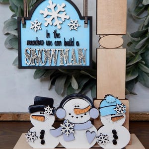 Puede incluir: Decoraci&oacute;n de invierno de madera con tres mu&ntilde;ecos de nieve y un letrero que dice "In the meadow we can build a SNOWMAN". Los mu&ntilde;ecos de nieve tienen sombreros negros, orejeras y gorros azules. El letrero tiene copos de nieve.