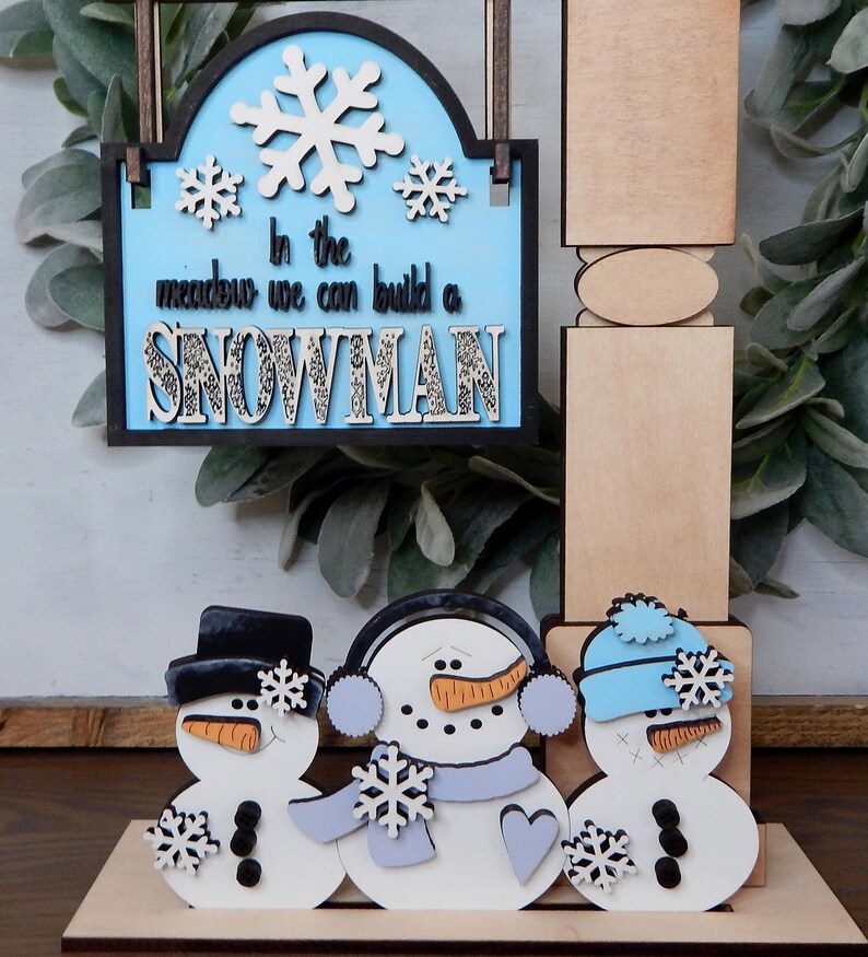 Puede incluir: Decoraci&oacute;n de invierno de madera con tres mu&ntilde;ecos de nieve y un letrero que dice "In the meadow we can build a SNOWMAN". Los mu&ntilde;ecos de nieve tienen sombreros negros, orejeras y gorros azules. El letrero tiene copos de nieve.