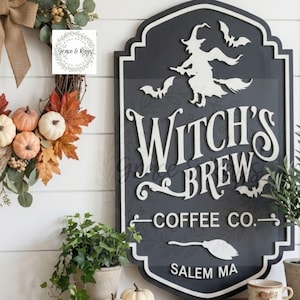 May include: A dark gray, shield-shaped wooden sign with white raised lettering that reads Witch's Brew Coffee Co. Salem MA. The sign features a white silhouette of a witch on a broomstick, three bats, and a broom icon, all with a white border.