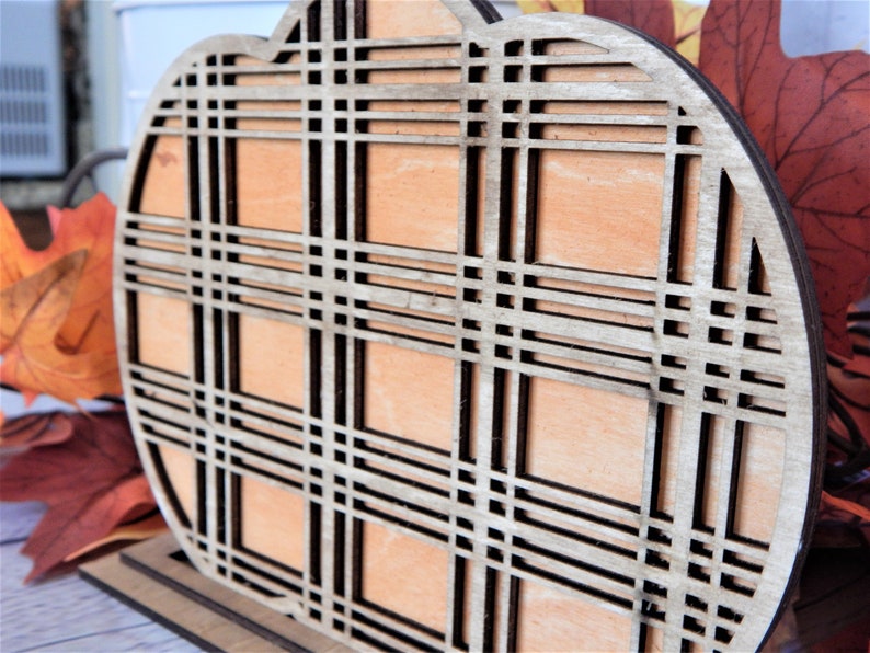 May include: A wooden pumpkin cutout with a plaid pattern. The pumpkin is painted a light brown color and has a dark brown plaid pattern.