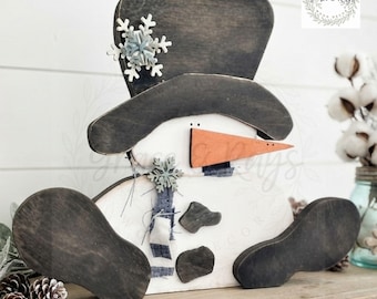 May include: A wooden snowman cutout featuring a white body, a dark gray top hat with a white snowflake accent, and a long orange carrot nose. The snowman has two dark gray buttons on its torso, a small blue fabric scarf, and two dark gray feet.