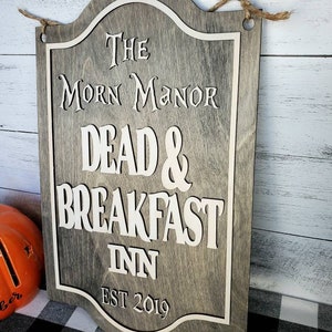 May include: A wooden sign with a gray background and white lettering. The sign reads "The Morn Manor Dead & Breakfast Inn Est 2019".