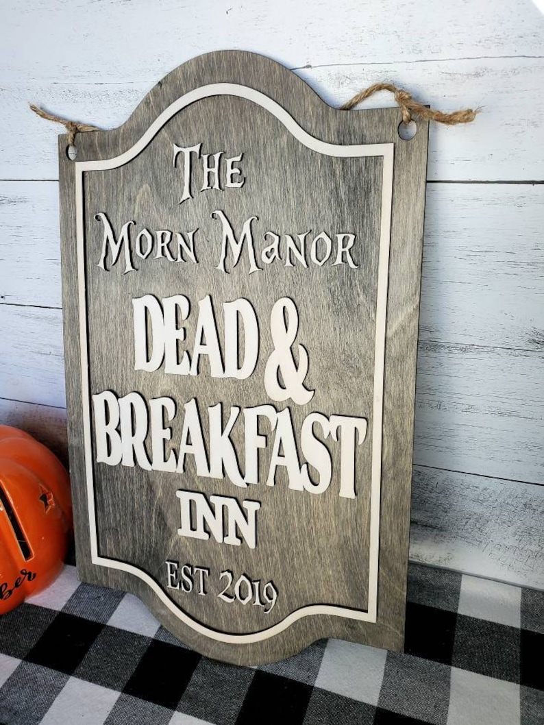 May include: A wooden sign with a gray background and white lettering. The sign reads "The Morn Manor Dead & Breakfast Inn Est 2019".