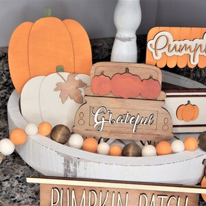 May include: A white wooden tray with a autumnal theme. The tray is decorated with orange and white pumpkins, a wooden truck with the word "Grateful" on it, and a sign that says "Pumpkins". There is also a white and orange beaded garland around the edge of the tray.