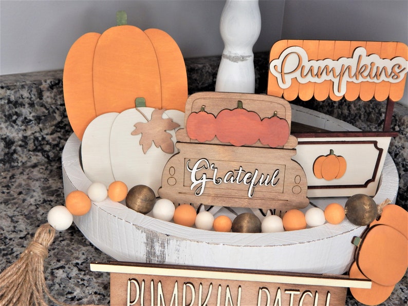 May include: A white wooden tray with a autumnal theme. The tray is decorated with orange and white pumpkins, a wooden truck with the word "Grateful" on it, and a sign that says "Pumpkins". There is also a white and orange beaded garland around the edge of the tray.
