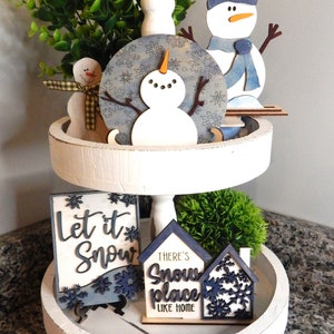 May include: A white tiered tray decorated with winter-themed wooden cutouts. The top tier features a snowman with a blue snowflake background, a snowman with a blue scarf, and a green bush. The bottom tier features a sign that says "Let it Snow" and a sign that says "There's no place like home".
