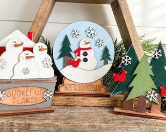 May include: Three wooden holiday tabletop decorations featuring a gray bucket labeled BUCKET OF FLAKES with three white snowmen, a circular sign labeled WINTER WONDERLAND with a snowman and trees, and a set of green pine trees with red birds.