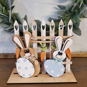 Pu&ograve; includere: Decorazione pasquale in legno con due coniglietti che tengono uova a pois, davanti a una staccionata con cuori. Carote e fiocchi aggiungono dettagli. La base &egrave; in legno chiaro.