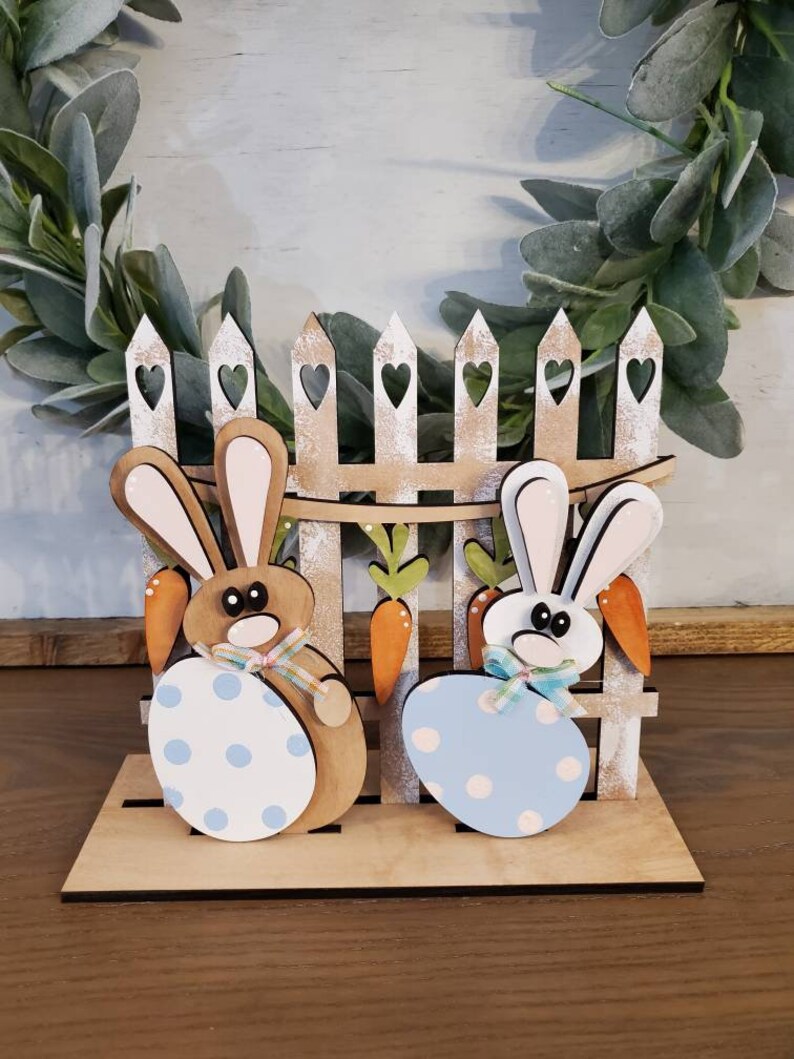 Pu&ograve; includere: Decorazione pasquale in legno con due coniglietti che tengono uova a pois, davanti a una staccionata con cuori. Carote e fiocchi aggiungono dettagli. La base &egrave; in legno chiaro.