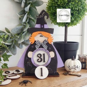 K&ouml;nnte beinhalten: Ein h&ouml;lzerner Halloween-Countdown-Kalender in Hexenform mit einem schwarzen Hut samt Fledermaus, orangefarbenen Haaren und einem violetten Umhang. In der Mitte steht der Text Countdown to Halloween mit der Zahl 31 und einer runden Holzscheibe.
