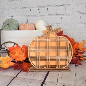 May include: A wooden pumpkin cutout with a plaid pattern, standing on a wooden base. The pumpkin is orange and the plaid pattern is brown and white. The pumpkin is surrounded by fall leaves.