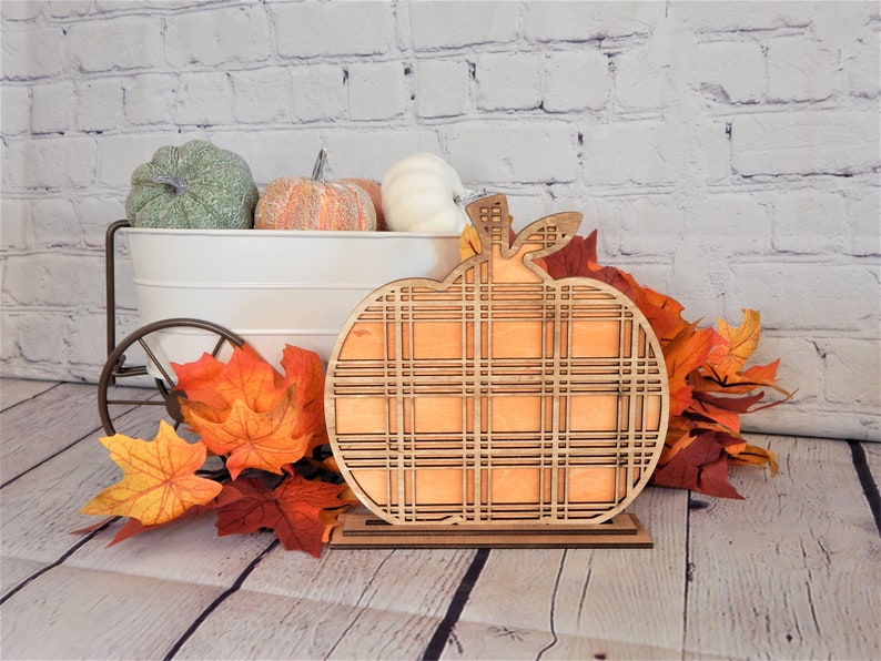 May include: A wooden pumpkin cutout with a plaid pattern, standing on a wooden base. The pumpkin is orange and the plaid pattern is brown and white. The pumpkin is surrounded by fall leaves.