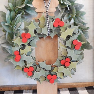 May include: A wooden Christmas wreath with green holly leaves and red berries. The wreath is mounted on a wooden frame and has a blank wooden tag in the center. A black and white checkered ribbon hangs from the top.