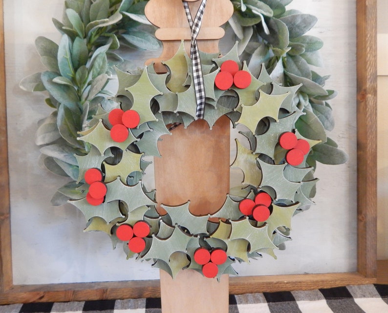 May include: A wooden Christmas wreath with green holly leaves and red berries. The wreath is mounted on a wooden frame and has a blank wooden tag in the center. A black and white checkered ribbon hangs from the top.