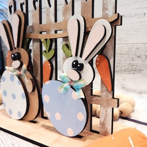 Pu&ograve; includere: Decorazione pasquale in legno con due coniglietti. Un coniglietto &egrave; marrone con un uovo bianco e blu a pois, l'altro &egrave; bianco con un uovo azzurro a pois. Carote e una staccionata in legno completano la scena.