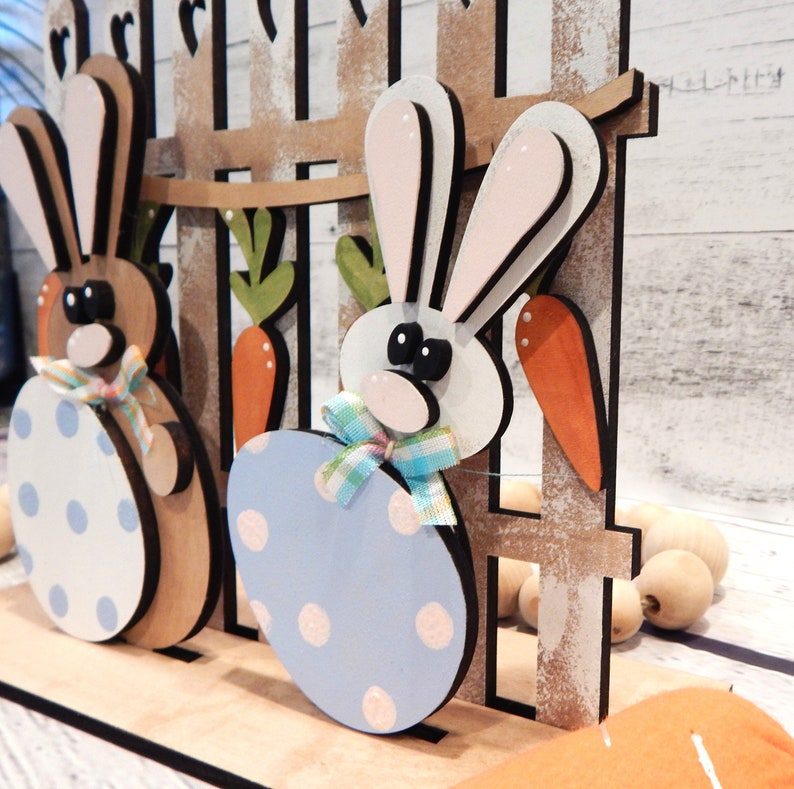 Pu&ograve; includere: Decorazione pasquale in legno con due coniglietti. Un coniglietto &egrave; marrone con un uovo bianco e blu a pois, l'altro &egrave; bianco con un uovo azzurro a pois. Carote e una staccionata in legno completano la scena.