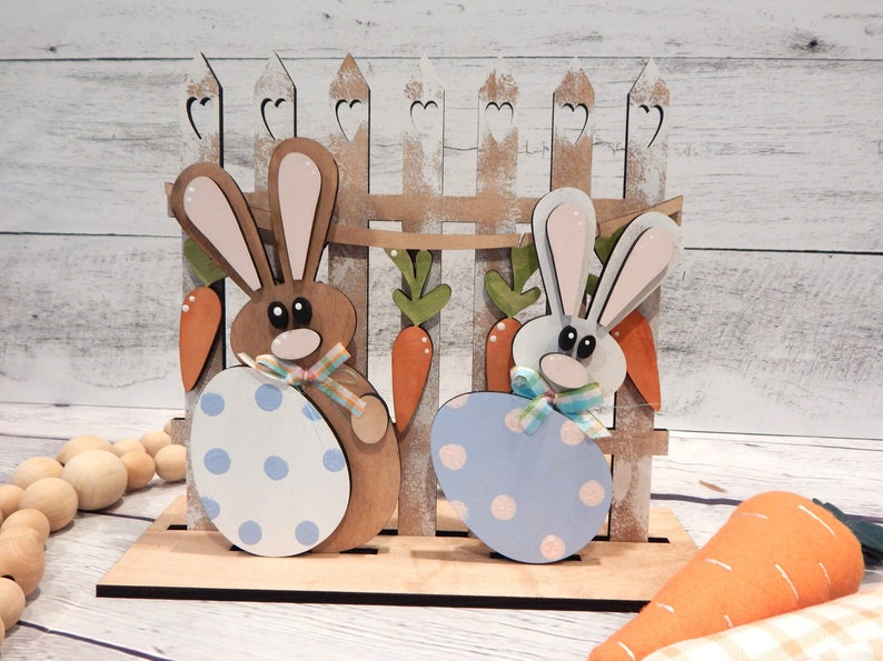 Pu&ograve; includere: Decorazione pasquale in legno con due coniglietti che tengono uova a pois, davanti a una staccionata con carote. I coniglietti sono marroni e bianchi con orecchie rosa, e le uova sono blu e bianche. Un'affascinante esposizione primaverile.