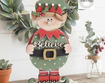 Puede incluir: Una figura de elfo de madera con un traje verde de lunares blancos, cuello rojo y cinturón negro con hebilla dorada. El elfo lleva un gorro verde con borde rojo y sostiene un cartel con la palabra Believe escrita en letras blancas.