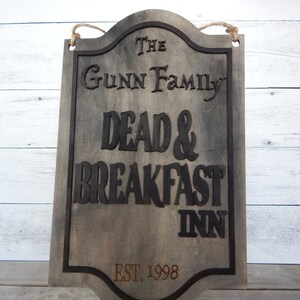 May include: A rustic wooden sign with black lettering that reads "The Gunn Family Dead & Breakfast Inn Est. 1998". The sign has a distressed, weathered look and is hanging from a rope.