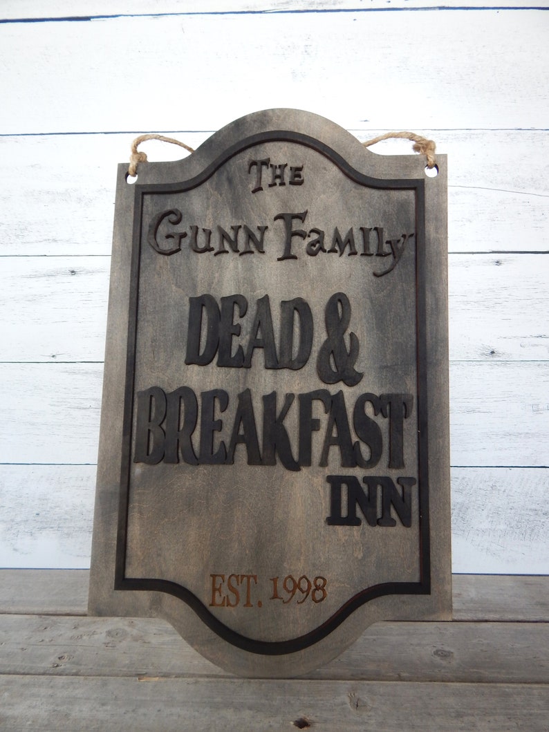 May include: A rustic wooden sign with black lettering that reads "The Gunn Family Dead & Breakfast Inn Est. 1998". The sign has a distressed, weathered look and is hanging from a rope.
