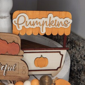 May include: A wooden pumpkin stand with a sign that reads "Pumpkins". The stand is painted white with brown trim and features a small pumpkin cutout on the front. The stand is displayed on a white tiered tray alongside a wooden truck and a beaded garland.