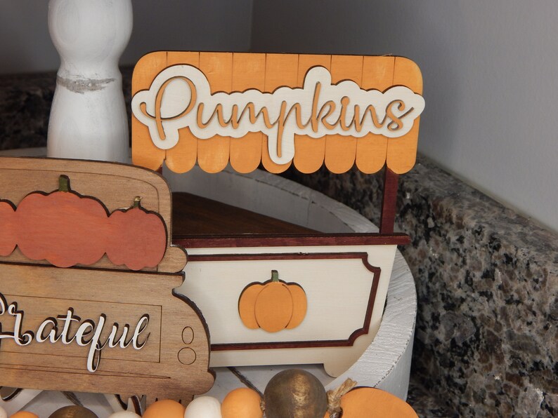 May include: A wooden pumpkin stand with a sign that reads "Pumpkins". The stand is painted white with brown trim and features a small pumpkin cutout on the front. The stand is displayed on a white tiered tray alongside a wooden truck and a beaded garland.