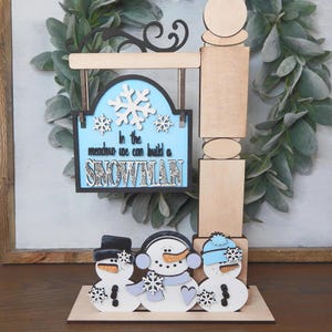 Puede incluir: Un letrero de madera con tem&aacute;tica invernal con tres figuras de mu&ntilde;ecos de nieve y el texto "In the meadow we can build a snowman". El letrero tiene un fondo azul claro con copos de nieve blancos. Los mu&ntilde;ecos de nieve tienen sombreros negros, bufandas y narices de zanahoria.