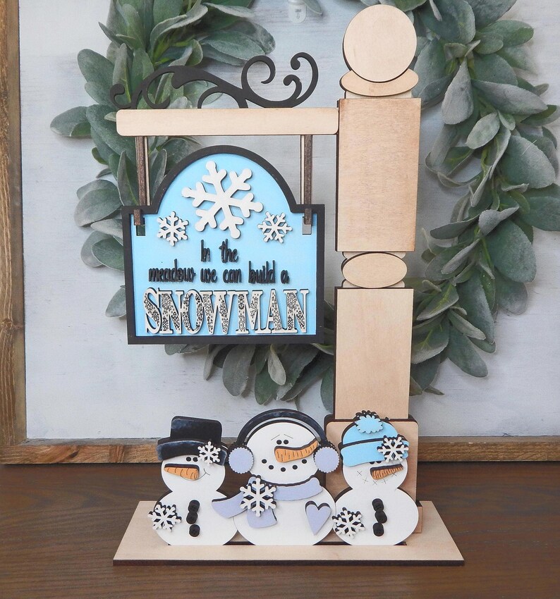 Puede incluir: Un letrero de madera con tem&aacute;tica invernal con tres figuras de mu&ntilde;ecos de nieve y el texto "In the meadow we can build a snowman". El letrero tiene un fondo azul claro con copos de nieve blancos. Los mu&ntilde;ecos de nieve tienen sombreros negros, bufandas y narices de zanahoria.