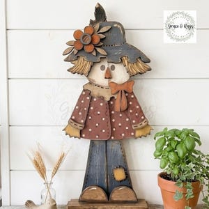 May include: A wooden scarecrow cutout featuring a dark hat with a large orange flower, a brown polka-dot jacket with a bow tie, and dark blue pants with a yellow patch. The figure has a light-colored face and straw-like hair accents.