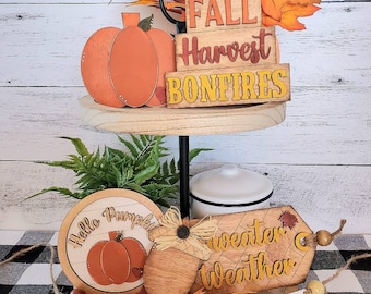Puede incluir: Una bandeja de dos niveles decorada con letreros de madera con temática otoñal, una calabaza y una pancarta. Los letreros dicen "Fall Harvest Bonfires", "Hello Pumpkin" y "Sweater Weather".