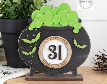 May include: A black wooden cauldron-shaped countdown sign featuring bright green bubbling liquid at the top, three small green bats, and a central circular dial displaying the number 31 with the text COUNTDOWN TO HALLOWEEN around the border.