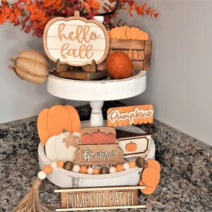 May include: A two-tiered white tray with a variety of autumn-themed decorations, including a wooden pumpkin sign that says "hello fall", a wooden crate with pumpkins, a wooden sign that says "pumpkins", a wooden wagon with the word "grateful" on it, and a wooden sign that says "pumpkin patch".