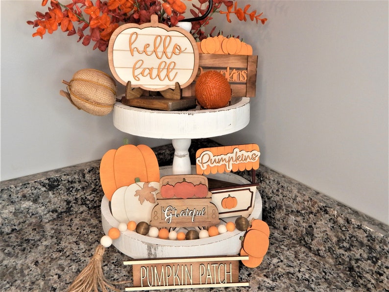 May include: A two-tiered white tray with a variety of autumn-themed decorations, including a wooden pumpkin sign that says "hello fall", a wooden crate with pumpkins, a wooden sign that says "pumpkins", a wooden wagon with the word "grateful" on it, and a wooden sign that says "pumpkin patch".