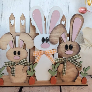 May include: Three wooden bunny napkin holders with pink ears and brown bodies. The bunnies are standing in front of a wooden fence with hearts cut out. The bunnies are wearing plaid bows and there are green leaves and orange carrots on the base.
