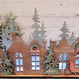 May include: Wooden Christmas village scene with three houses, a sleigh, and trees. The houses have windows and wreaths with red and white ribbon. The trees are green and have snowflakes. The sleigh is red and has a white trim.