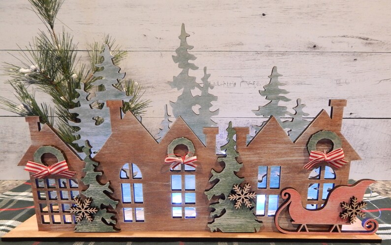 May include: Wooden Christmas village scene with three houses, a sleigh, and trees. The houses have windows and wreaths with red and white ribbon. The trees are green and have snowflakes. The sleigh is red and has a white trim.