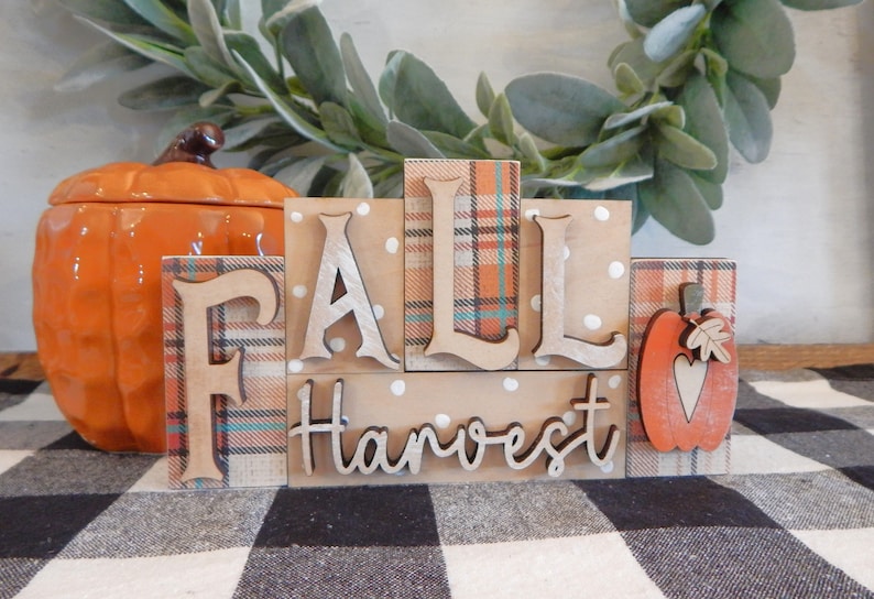 May include: Wooden block sign with the words 'Fall Harvest' in a rustic style. The blocks are decorated with plaid and polka dot patterns. A small wooden pumpkin with a heart is on the right side of the sign.