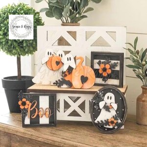 Puede incluir: Un conjunto de decoraci&oacute;n para bandeja escalonada con figuras de madera de fantasmas y calabazas, un letrero cuadrado con rayas blancas y negras y flores naranjas, un letrero con la palabra Boo y un &oacute;valo con un fantasma y flores.