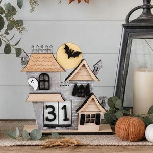 Puede incluir: Un calendario de casa de Halloween de madera con un dise&ntilde;o de murci&eacute;lago y luna. El calendario muestra el n&uacute;mero 31. La casa tiene un fantasma y varios detalles arquitect&oacute;nicos. Una calabaza y una vela est&aacute;n en el fondo.