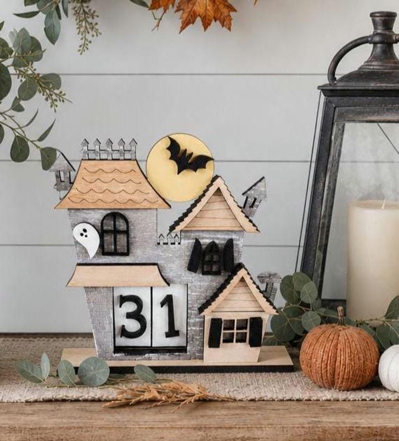 Puede incluir: Un calendario de casa de Halloween de madera con un dise&ntilde;o de murci&eacute;lago y luna. El calendario muestra el n&uacute;mero 31. La casa tiene un fantasma y varios detalles arquitect&oacute;nicos. Una calabaza y una vela est&aacute;n en el fondo.