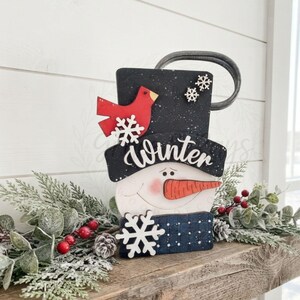 May include: A decorative snowman sign featuring a black top hat with white speckles, a red cardinal, and two small white snowflakes. The word Winter is written in white script across the hat brim. A large white snowflake sits below the orange nose.