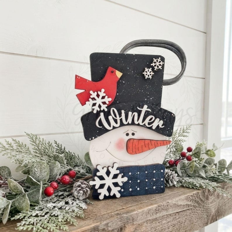May include: A decorative snowman sign featuring a black top hat with white speckles, a red cardinal, and two small white snowflakes. The word Winter is written in white script across the hat brim. A large white snowflake sits below the orange nose.