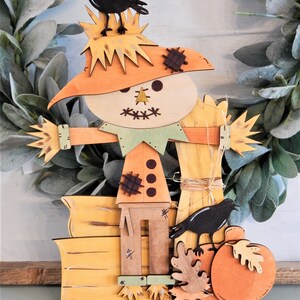 May include: Wooden fall-themed decoration featuring a scarecrow with a straw hat, a green collar, and a pumpkin. Two black crows are perched on the scarecrow and pumpkin. The decoration is painted in shades of orange, yellow, and brown.
