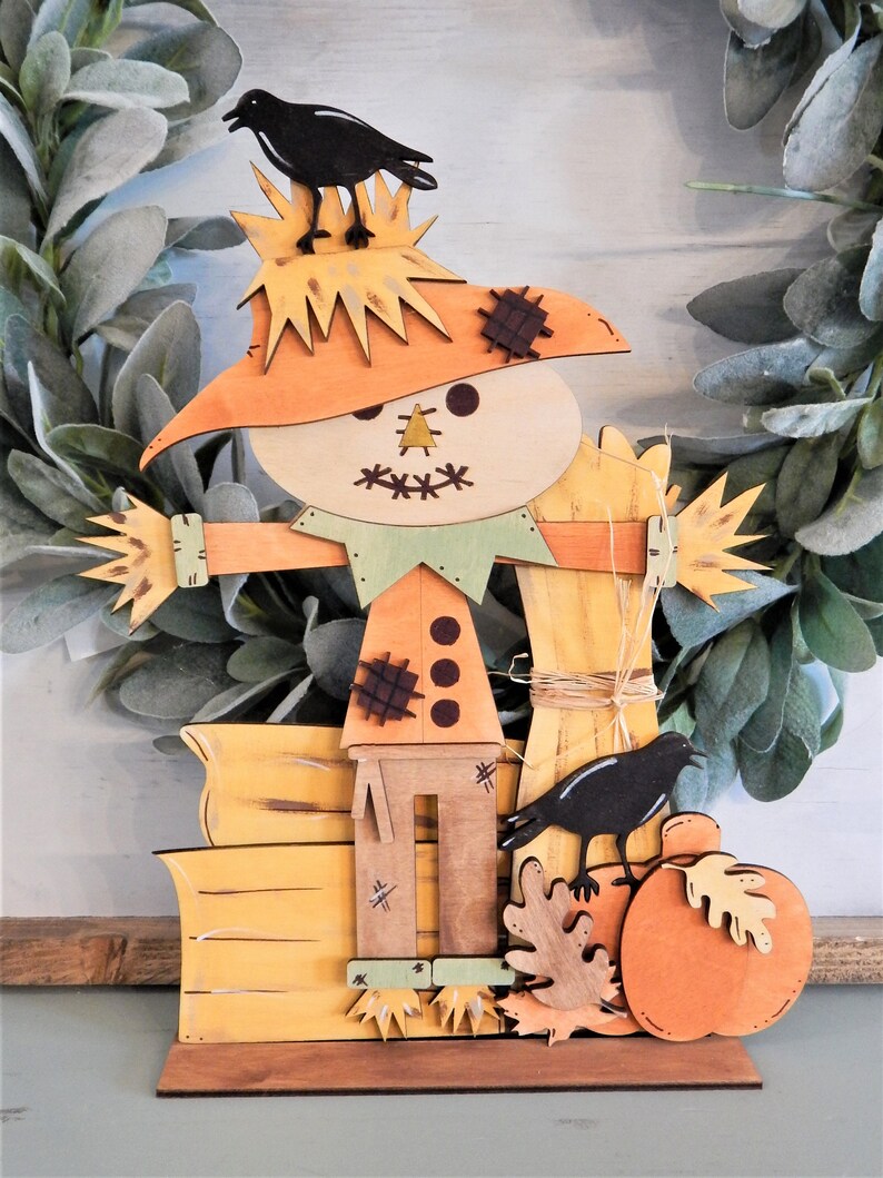 May include: Wooden fall-themed decoration featuring a scarecrow with a straw hat, a green collar, and a pumpkin. Two black crows are perched on the scarecrow and pumpkin. The decoration is painted in shades of orange, yellow, and brown.