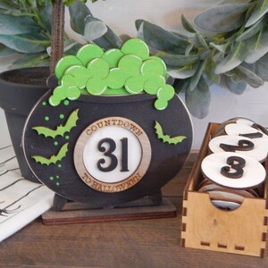 Pu&ograve; includere: Un calendario a tema Halloween con un calderone nero con pozione verde gorgogliante e ritagli di pipistrelli. Il calendario mostra il numero 31 e include una scatola di legno con dischi numerati per il conto alla rovescia quotidiano.
