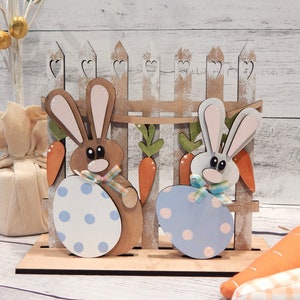 Pu&ograve; includere: Decorazione pasquale in legno con due coniglietti che tengono uova a pois, davanti a una staccionata con carote. I coniglietti sono marroni e bianchi, con uova dai colori pastello. Un'esposizione primaverile festosa.