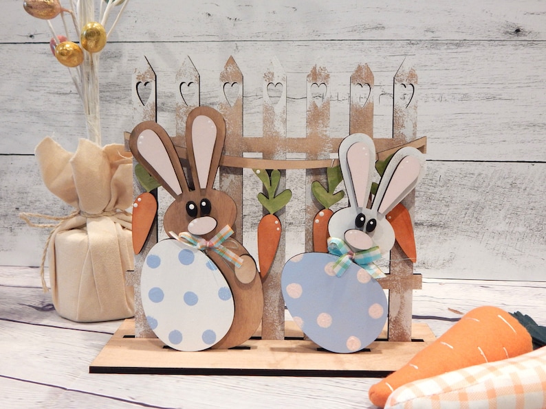 Pu&ograve; includere: Decorazione pasquale in legno con due coniglietti che tengono uova a pois, davanti a una staccionata con carote. I coniglietti sono marroni e bianchi, con uova dai colori pastello. Un'esposizione primaverile festosa.