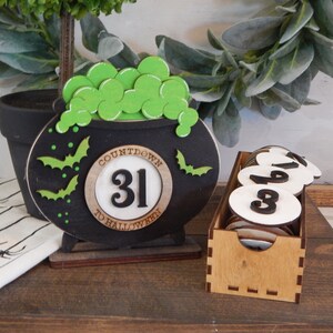 Pu&ograve; includere: Decorazione a forma di calderone nero per il conto alla rovescia di Halloween con pozione verde gorgogliante e pipistrelli. Il testo recita "Countdown to Halloween 31". Include una scatola di legno con dischi numerati.
