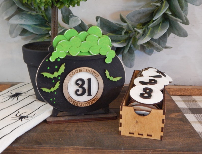 Pu&ograve; includere: Decorazione a forma di calderone nero per il conto alla rovescia di Halloween con pozione verde gorgogliante e pipistrelli. Il testo recita "Countdown to Halloween 31". Include una scatola di legno con dischi numerati.
