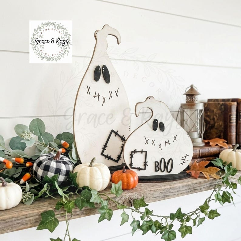 May include: Two white wooden ghost decorations with black stitched-style eyes, mouths, and patches. The smaller ghost features the word BOO in black lettering. They are displayed alongside small pumpkins, greenery, and a metal lantern.